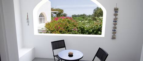 A room with a white interior, a round table with a stone centerpiece, and two black chairs. A large arched window reveals a garden with pink flowers and green foliage.