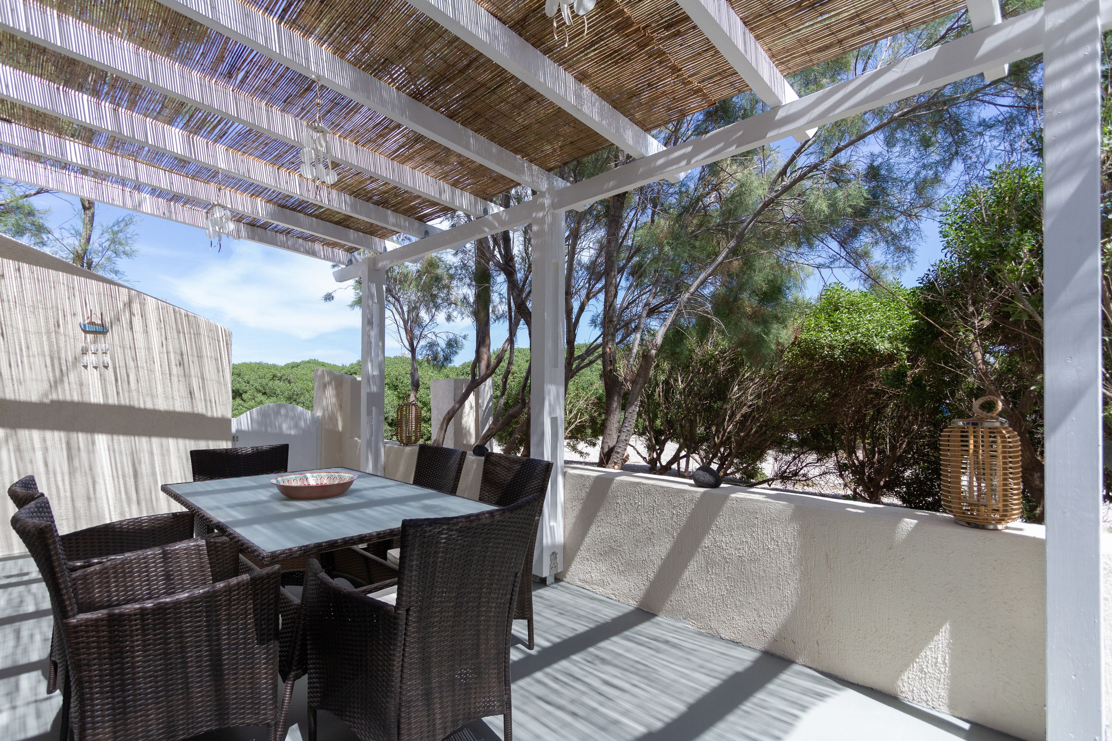 Un patio exterior con una mesa y sillas, rodeado por una pérgola blanca y vistas a árboles y colinas.