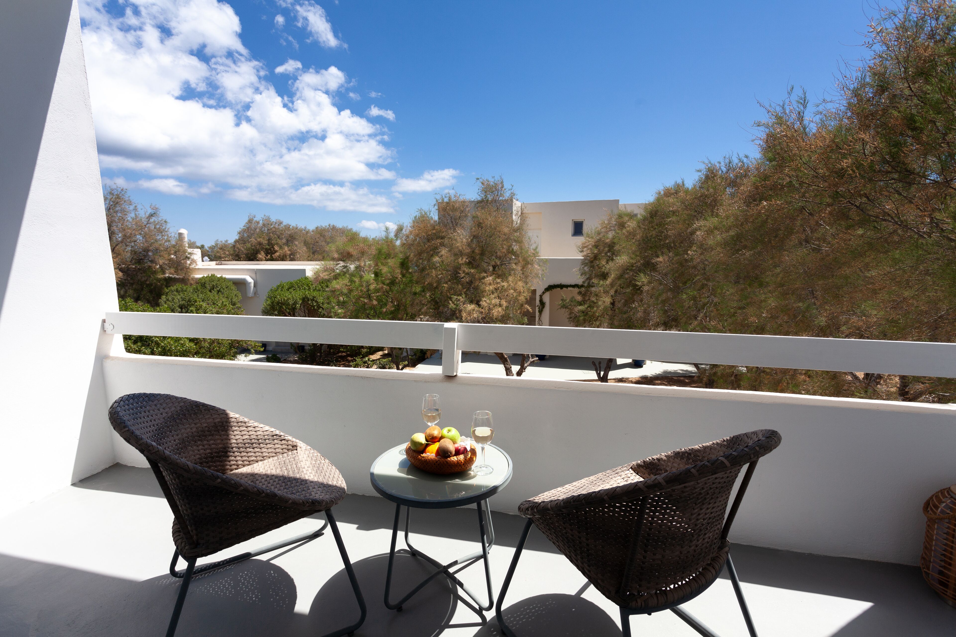 Un balcón con dos sillones de mimbre, una pequeña mesa redonda con un frutero y dos copas de vino, y vistas a árboles y edificios.