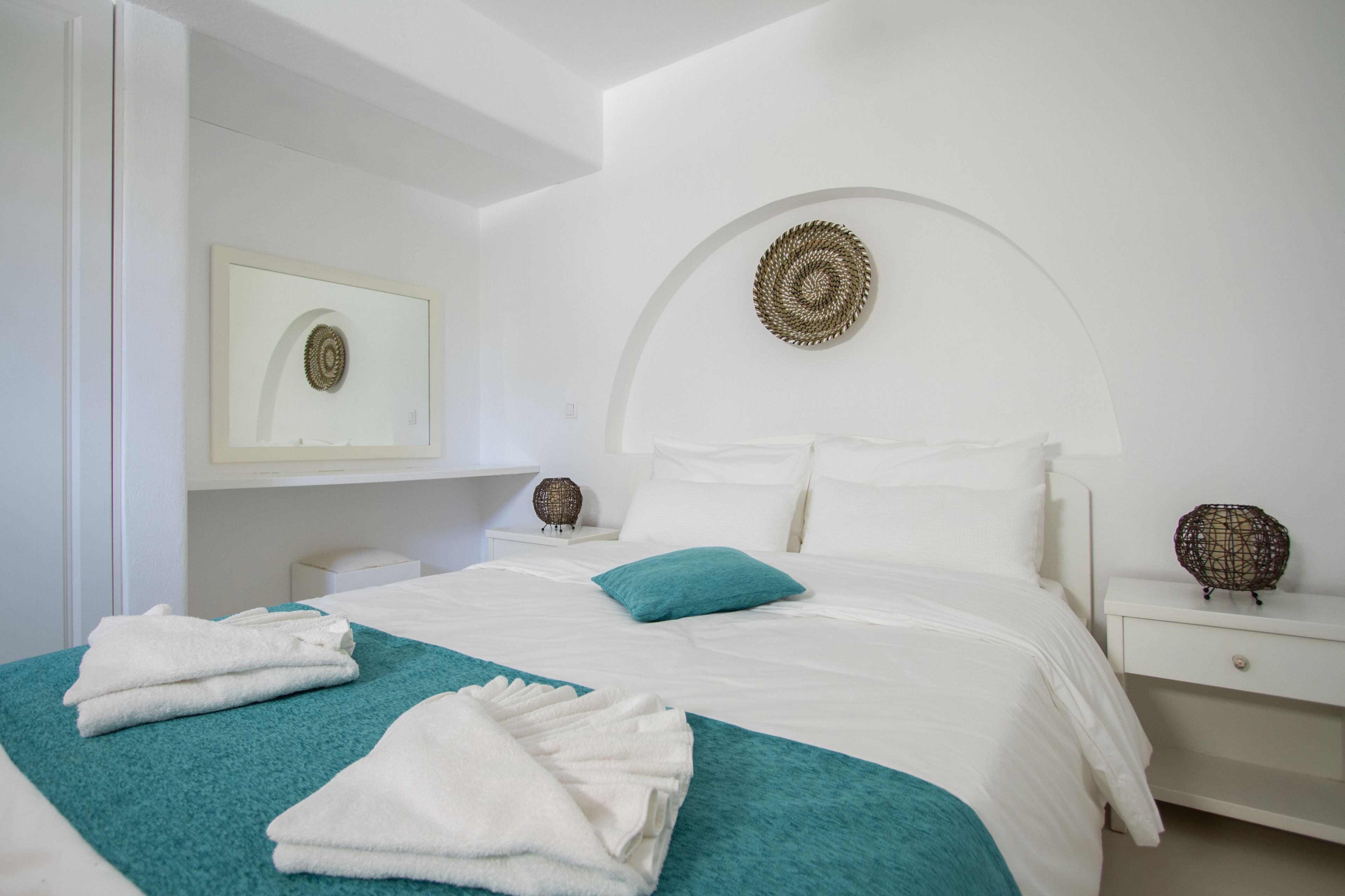 Cama bem arrumada com lençóis brancos e detalhes verde-água, mesa de cabeceira com um objeto decorativo e um espelho fixado na parede.