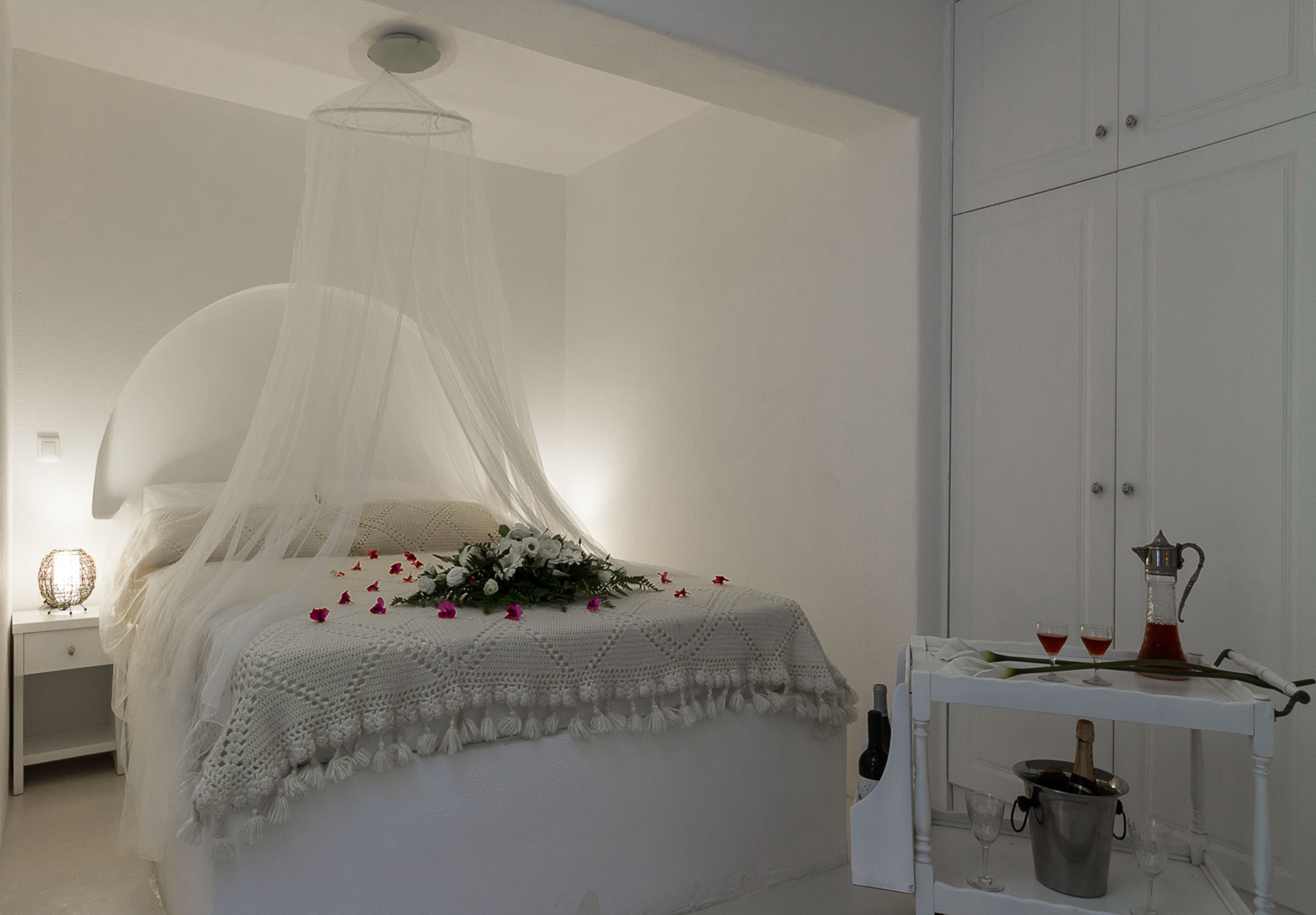 A bedroom with a canopy bed, a bedside table with a lamp, and a small table with a bottle and glasses.
