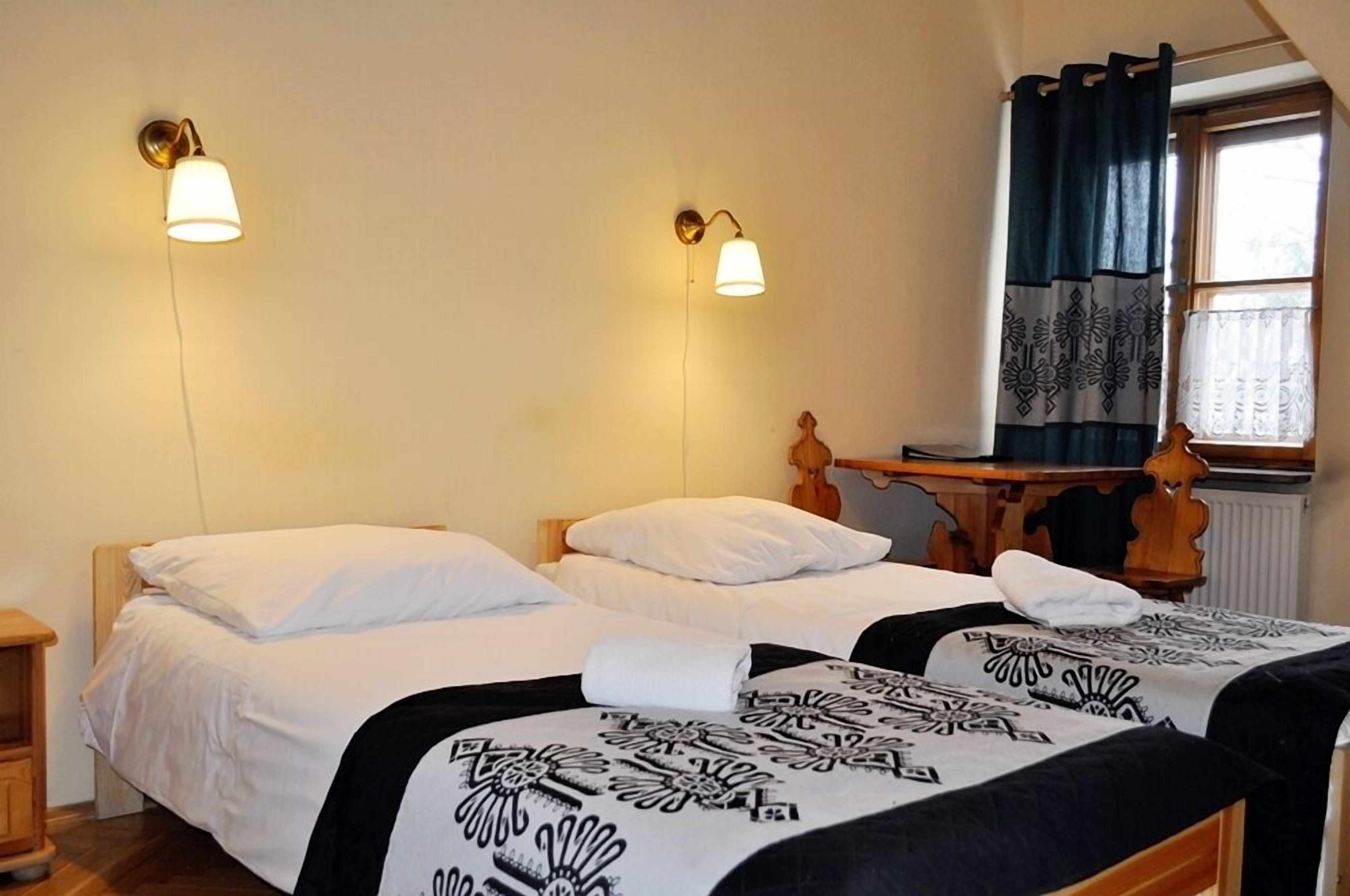 Una habitació amb dos llits, cadascun amb roba de llit blanca i mantilles de colors negres i blancs. Hi ha làmpades murals a cada costat dels llits. Un escriptori de fusta amb una cadira està situat a prop d'una finestra amb cortines estampades.
