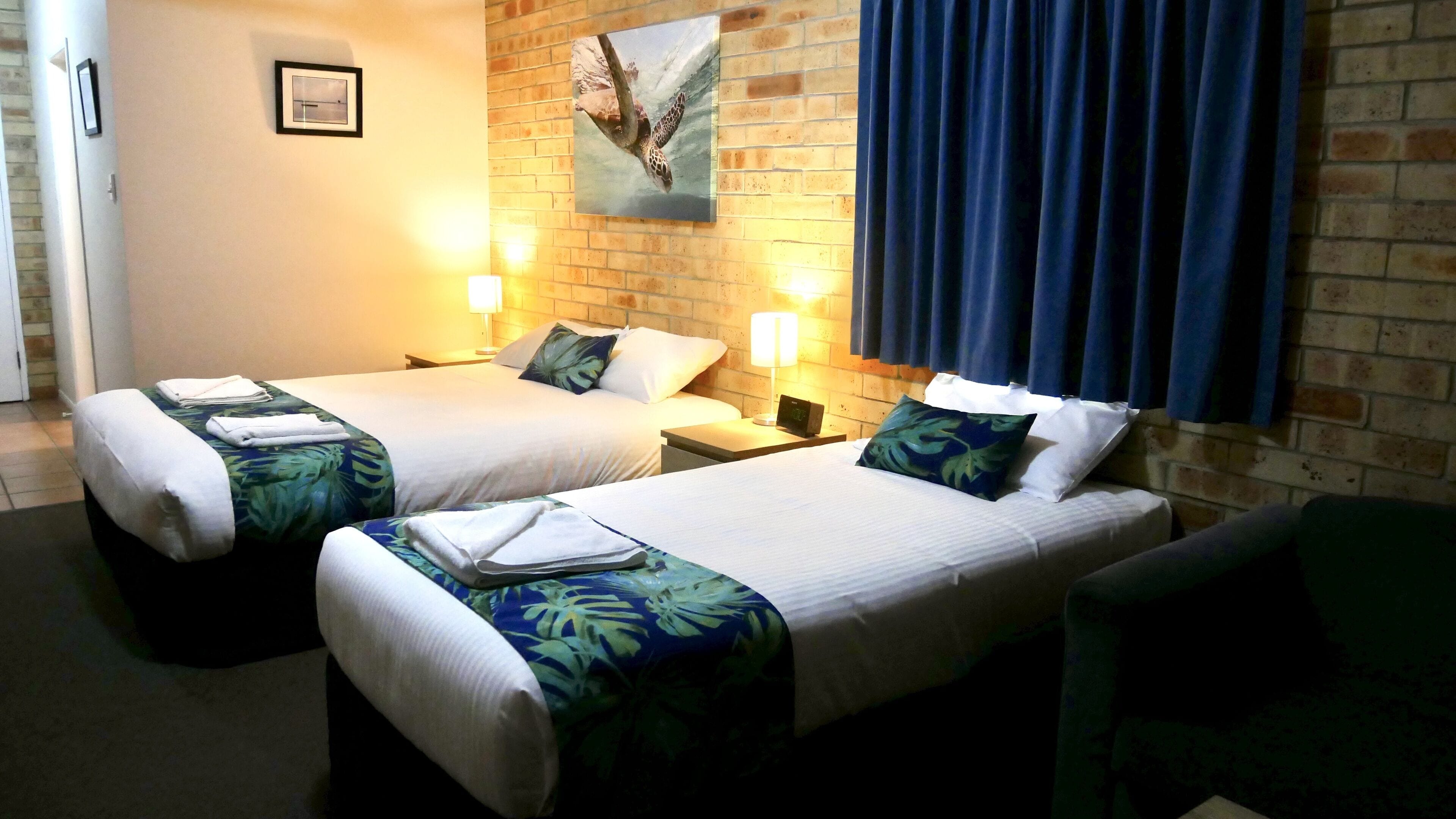 Une chambre d’hôtel avec deux lits, chacun équipé d’un oreiller et d’une couverture, une table de chevet avec une lampe et un tableau accroché au mur.