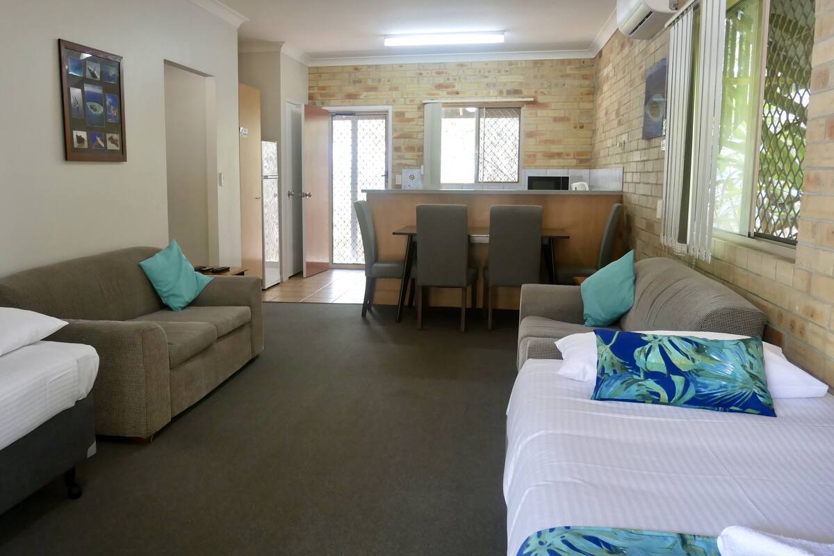 A hotel room with two beds, a sofa, a dining table with chairs, and a kitchen area.