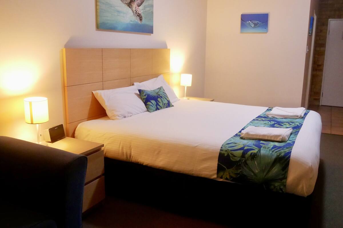 A hotel room with a bed, bedside table, lamp, and a painting on the wall.