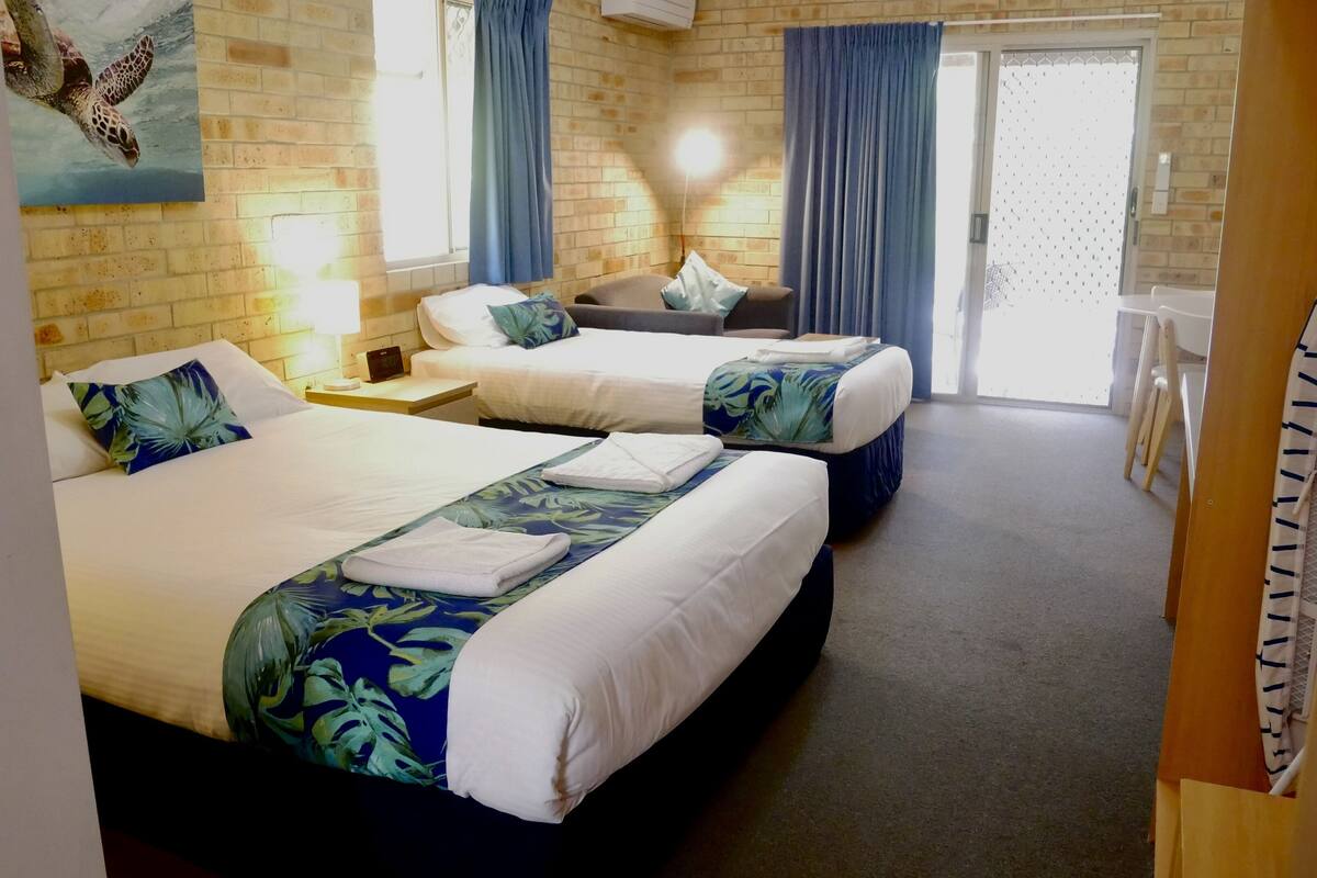 A hotel room with two beds, a small table, a chair, a window with curtains, and a wall-mounted air conditioner.