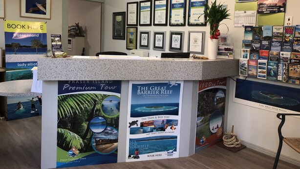 A reception desk with framed certificates, a 'Book Here' sign, and promotional posters for tours and resorts. - Emeraldene Inn & Eco-Lodge (Urraween)