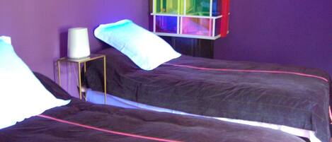 A room with two beds, a small table with a lamp, and a colorful storage unit against the wall.