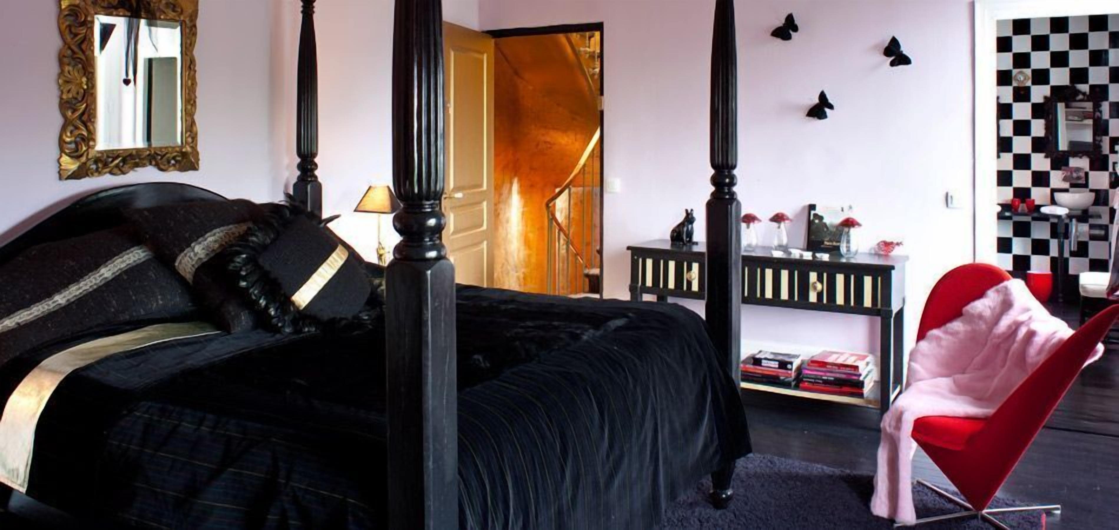 A bedroom with a four-poster bed, a red chair, a console table with books, and a checkered wall pattern.