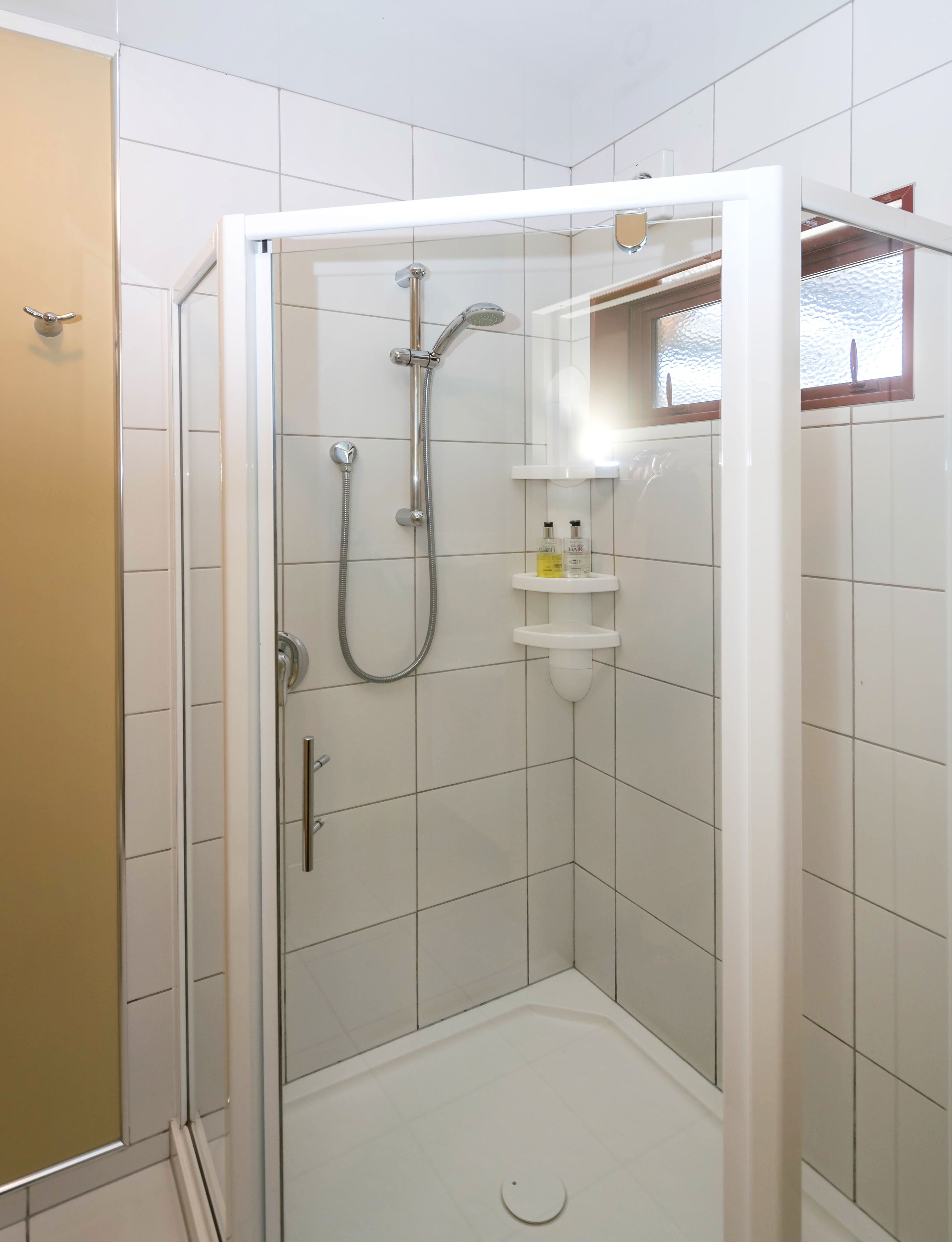 Ducha a pie con mampara de vidrio, paredes de azulejos blancos y un estante pequeño con artículos de aseo.