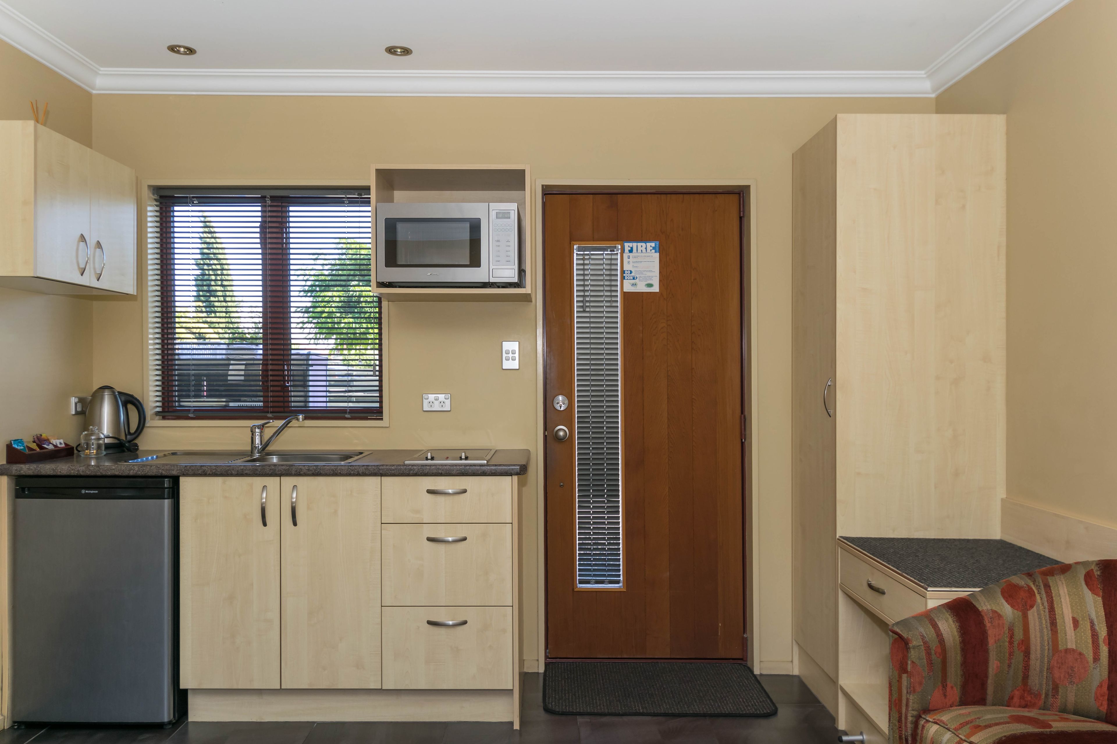 A compact kitchen with a microwave, sink, and dishwasher. There is a door with a vent and a small mat at the entrance.