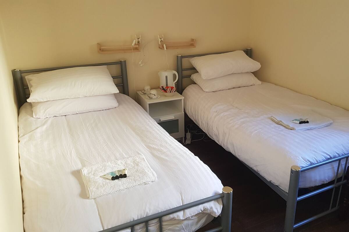 Two single beds with white bedding, a bedside table with a kettle and cups, and a wooden shelf above the beds.