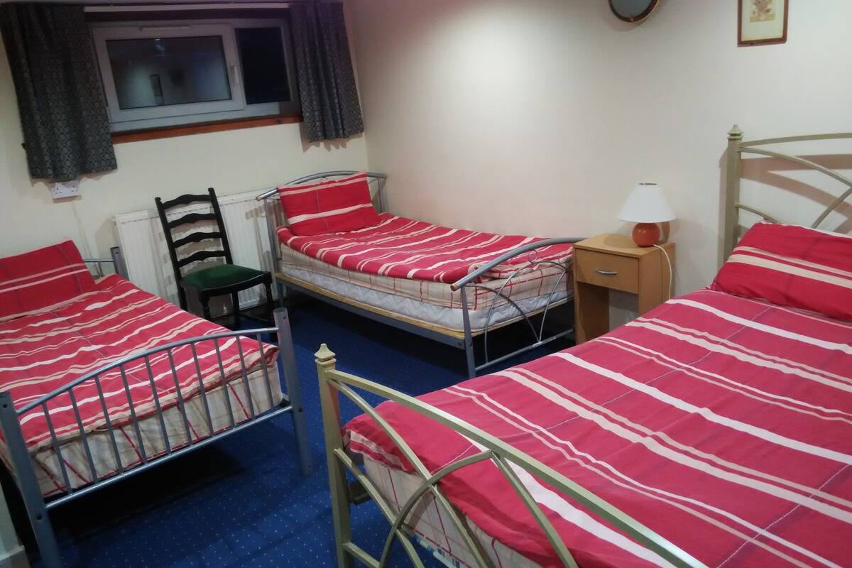 A room with two metal bunk beds, a single bed, a chair, a bedside table with a lamp, a window with curtains, and a radiator.