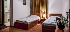 Camera d'albergo con due letti singoli, una testiera in legno, un comodino con una lampada, una pianta in vaso e un bagno visibile attraverso una porta aperta.