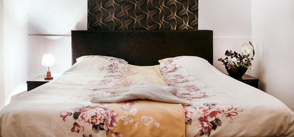 Un letto ben rifatto con biancheria da letto floreale, una testiera scura e un comodino con una lampada e un vaso di fiori.