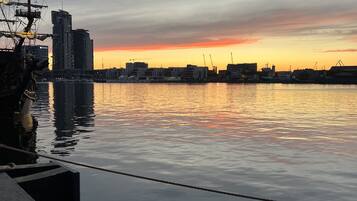 Vista al agua con un río tranquilo, un muelle con un barco y el horizonte de una ciudad al atardecer.