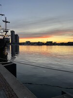 Vista al agua con un río tranquilo, un muelle con un barco y el horizonte de una ciudad al atardecer.