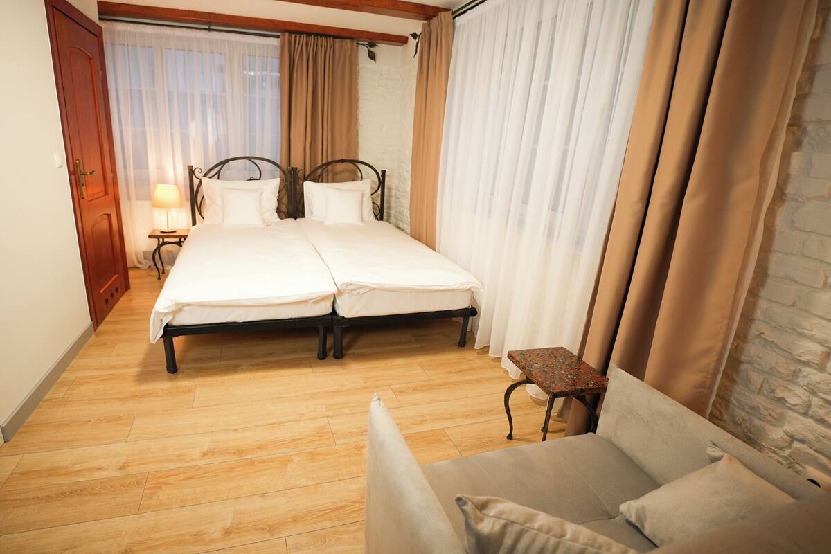 A hotel room with a double bed, a small table, a sofa, and a window with curtains.