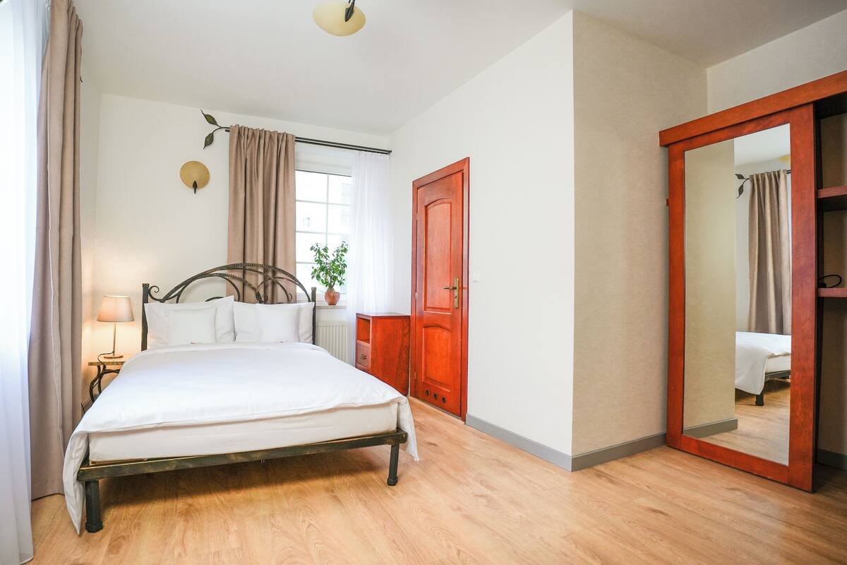 A bedroom with a bed, a nightstand, a lamp, a window with curtains, a wooden door, and a mirror.