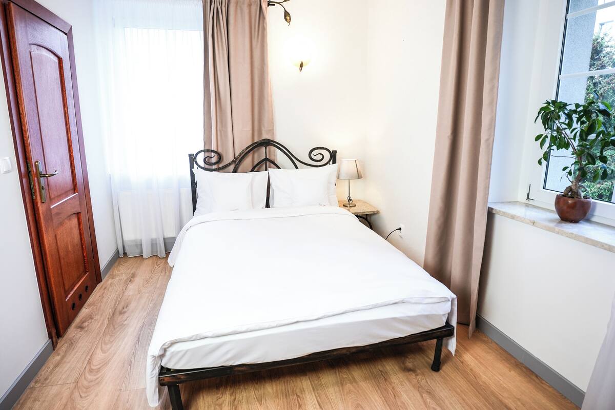 A neatly made bed in a room with a wooden floor, a window with a plant, and a door.