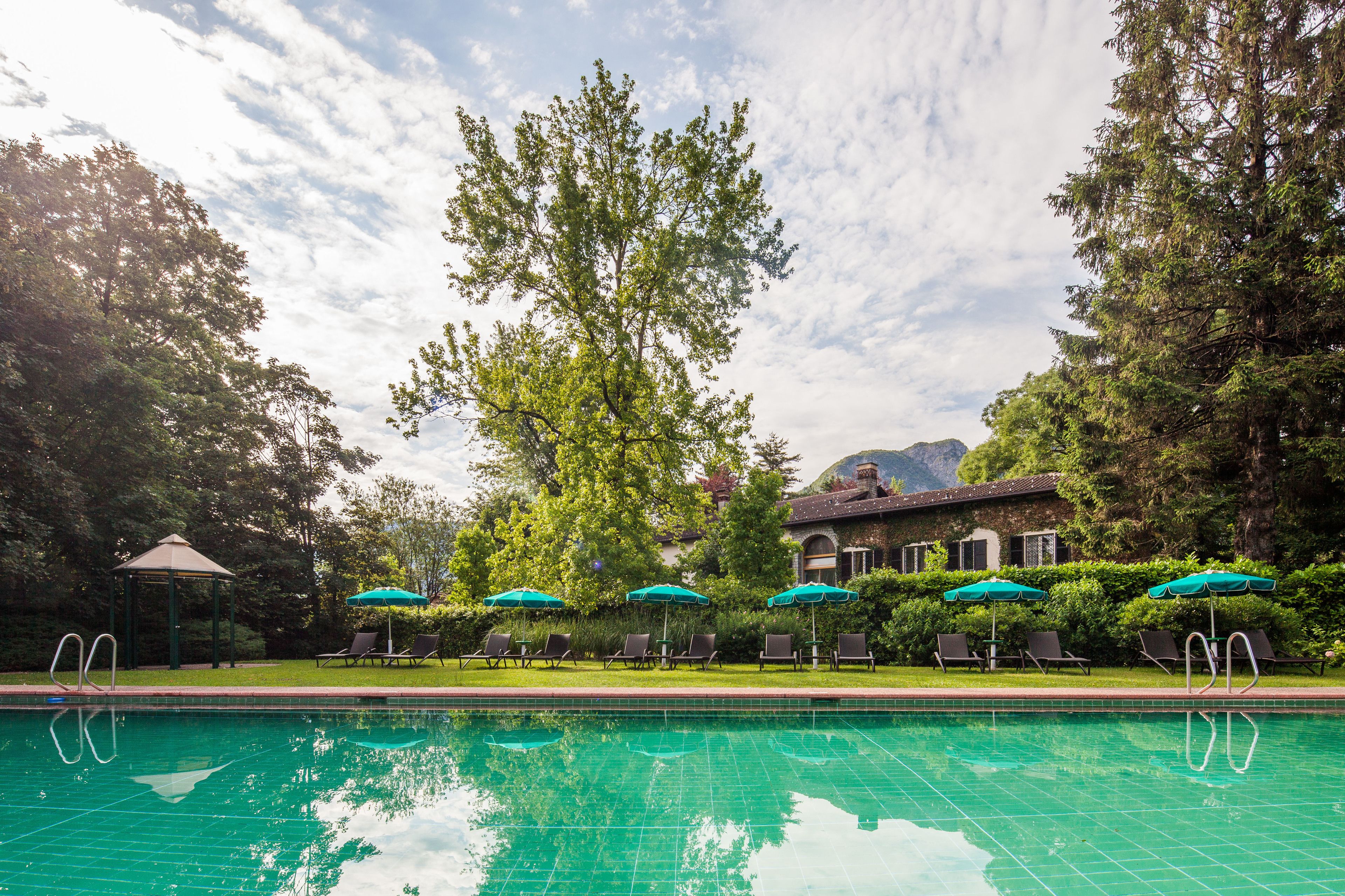 A swimming pool with lounge chairs and umbrellas, surrounded by trees and a building with a mountain in the background.