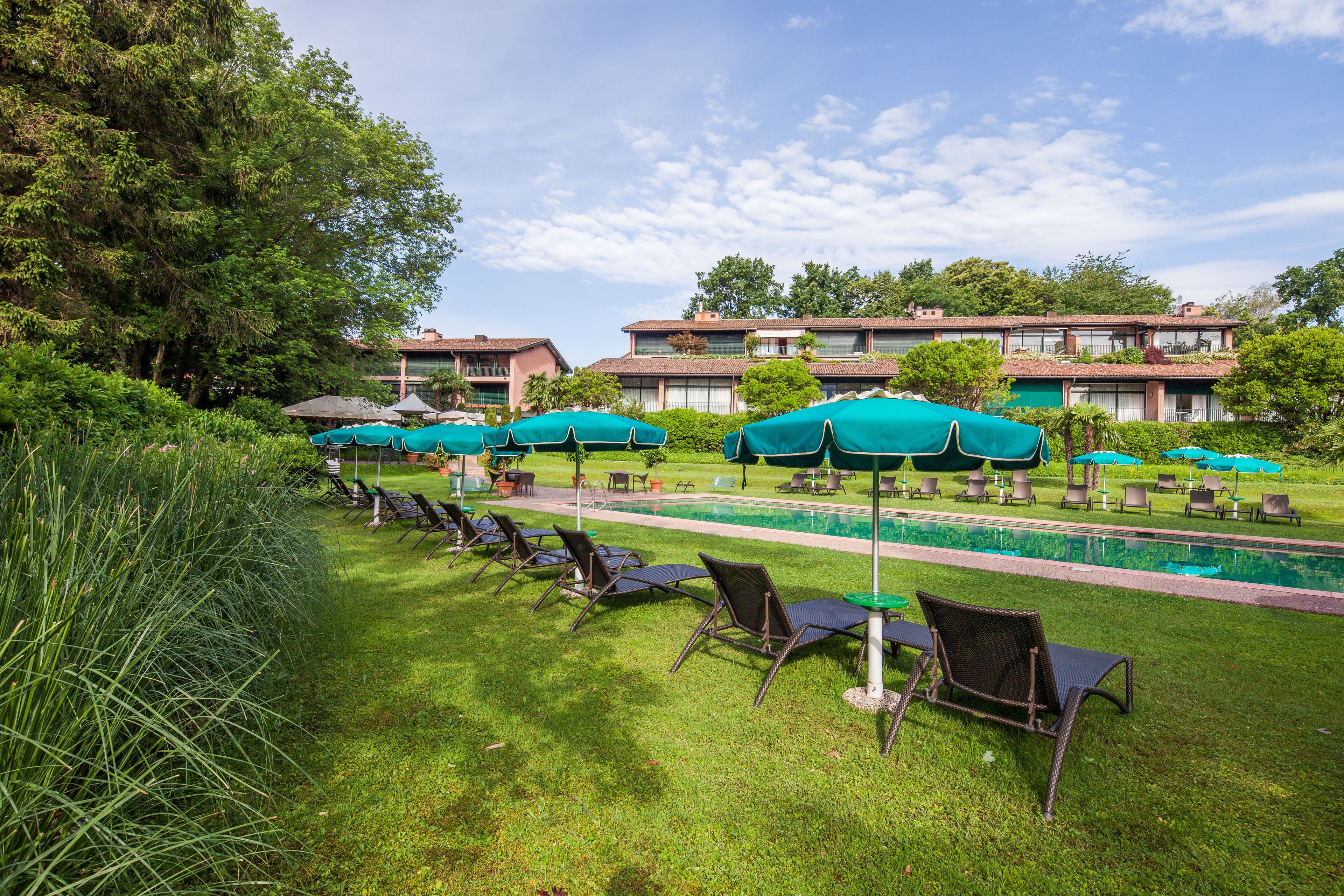 Un espace aménagé au bord de la piscine, avec des chaises longues et des parasols, entouré de verdure et avec des bâtiments en arrière-plan.