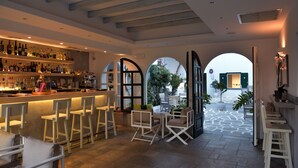A bar area with white bar stools and a variety of bottles on shelves. There are arched doorways leading to outdoor seating. - Kalypso Hotel (Paros)