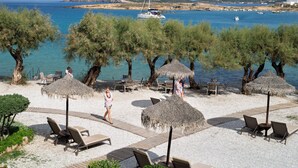 A beachside area with lounge chairs, umbrellas, and a view of the sea and boats. - Kalypso Hotel (Paros)