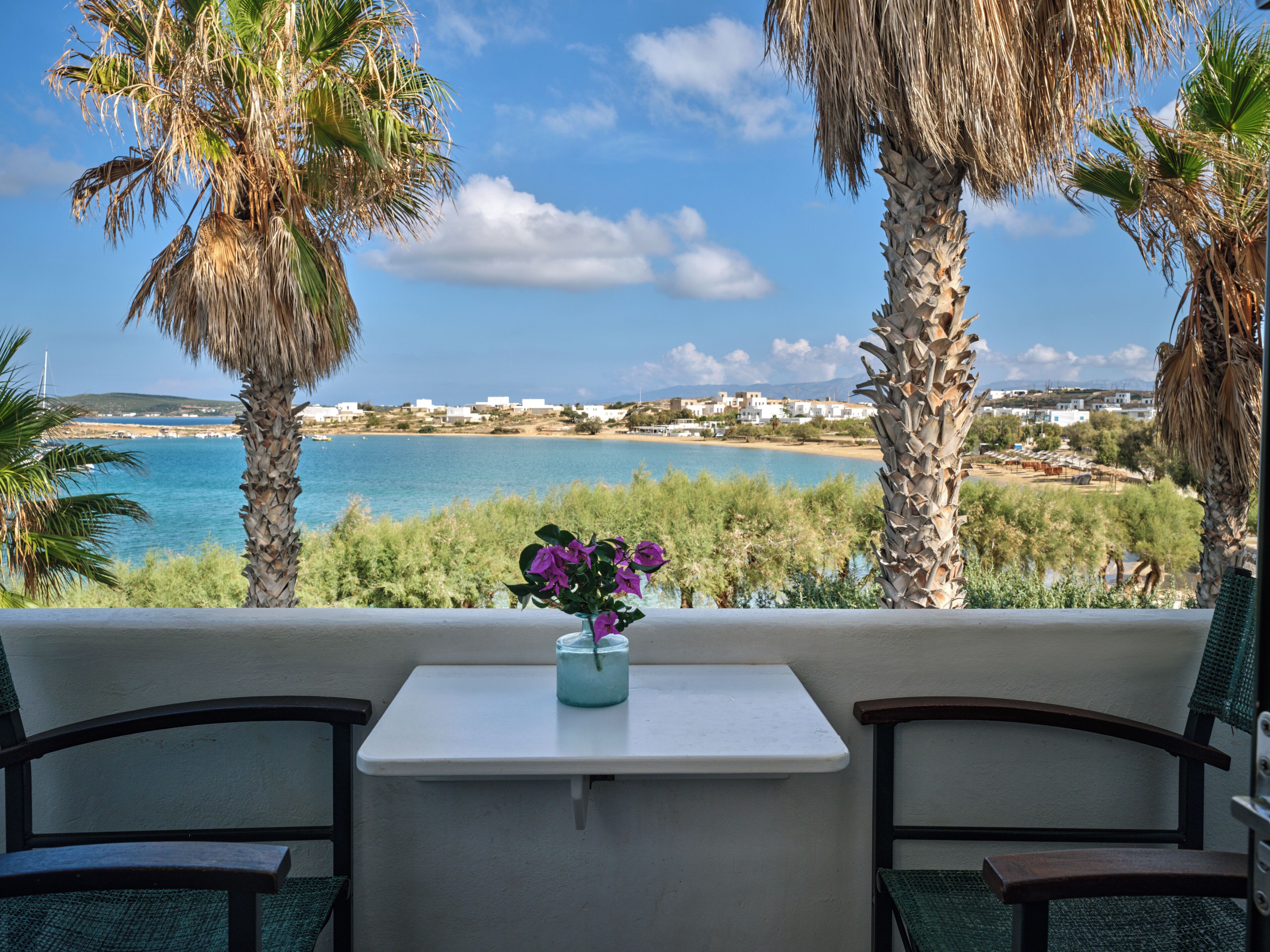 Un balcón con una mesa blanca y dos sillas, con vistas a un paisaje costero con palmeras y una vista del mar y edificios al fondo.