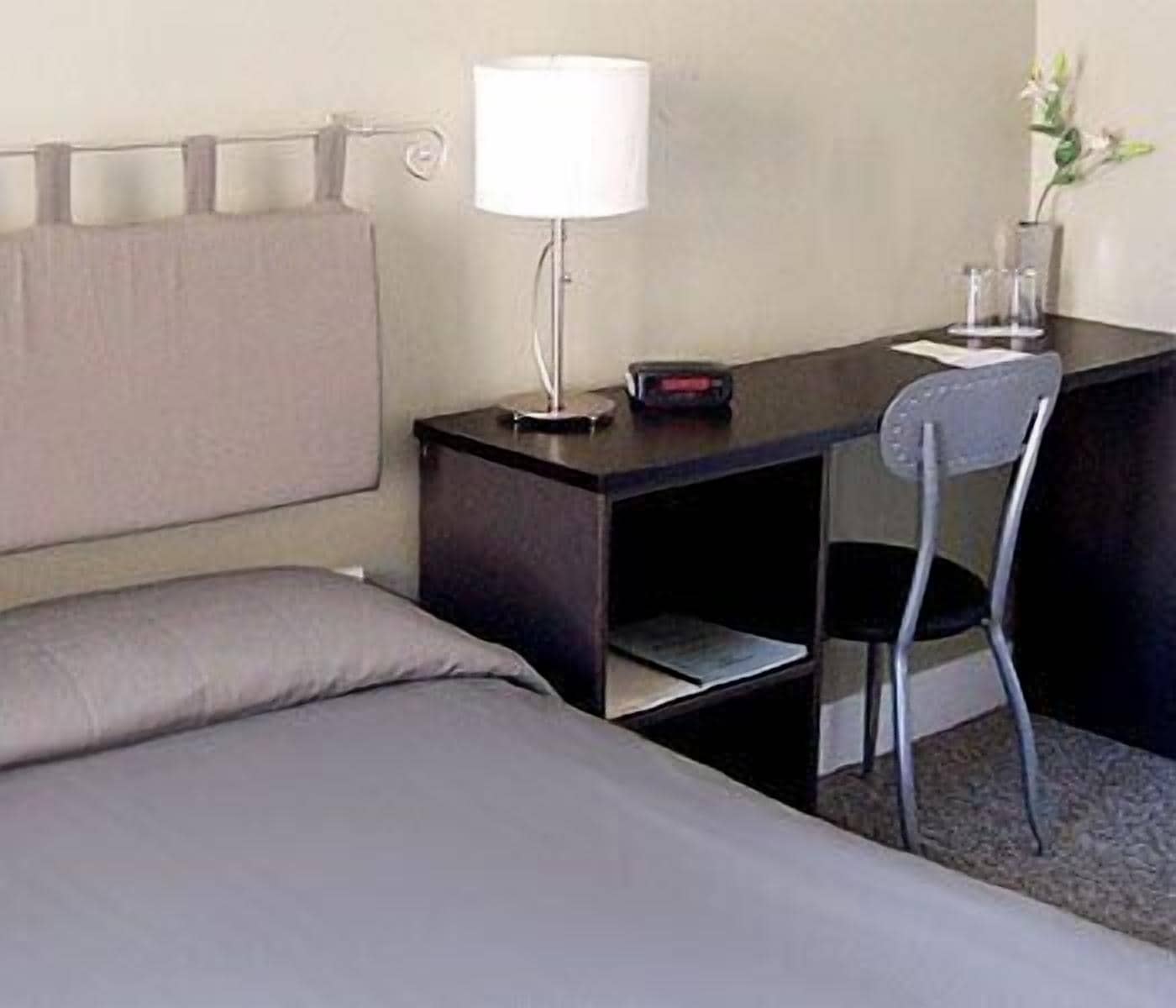 Ein Hotelzimmer mit Bett, Schreibtisch mit Lampe, Stuhl und einer Vase mit Blumen.