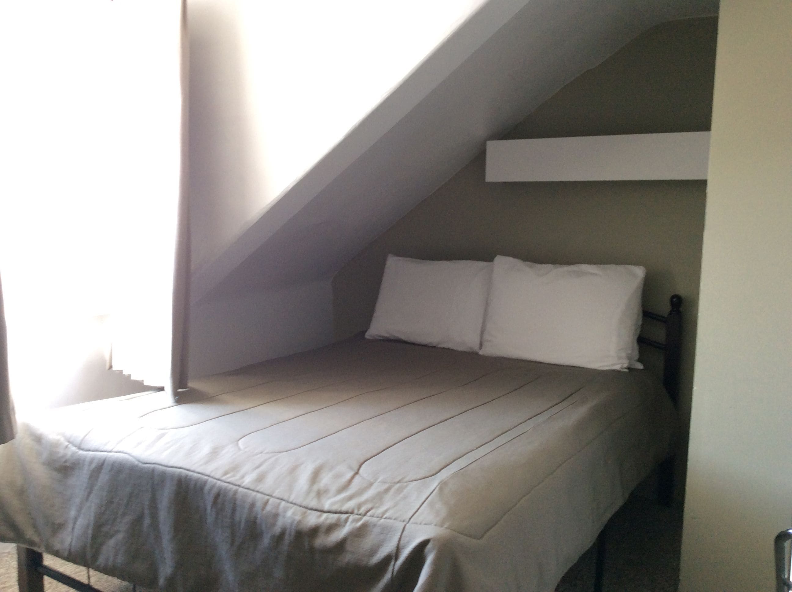 Une petite chambre avec un lit simple, un plafond en pente et une fenêtre sur le côté gauche.