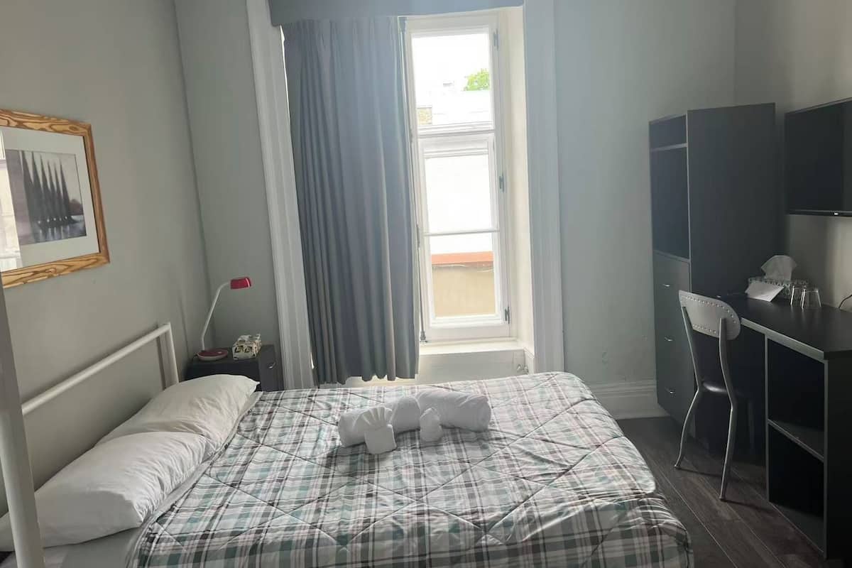 A hotel room with a bed, a desk, a chair, a window with curtains, a bedside table with a lamp, and a framed picture on the wall.