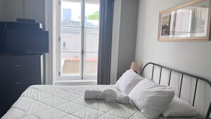 Quarto de hotel com uma cama, cômoda, janela com cortinas, luminária e um quadro na parede.