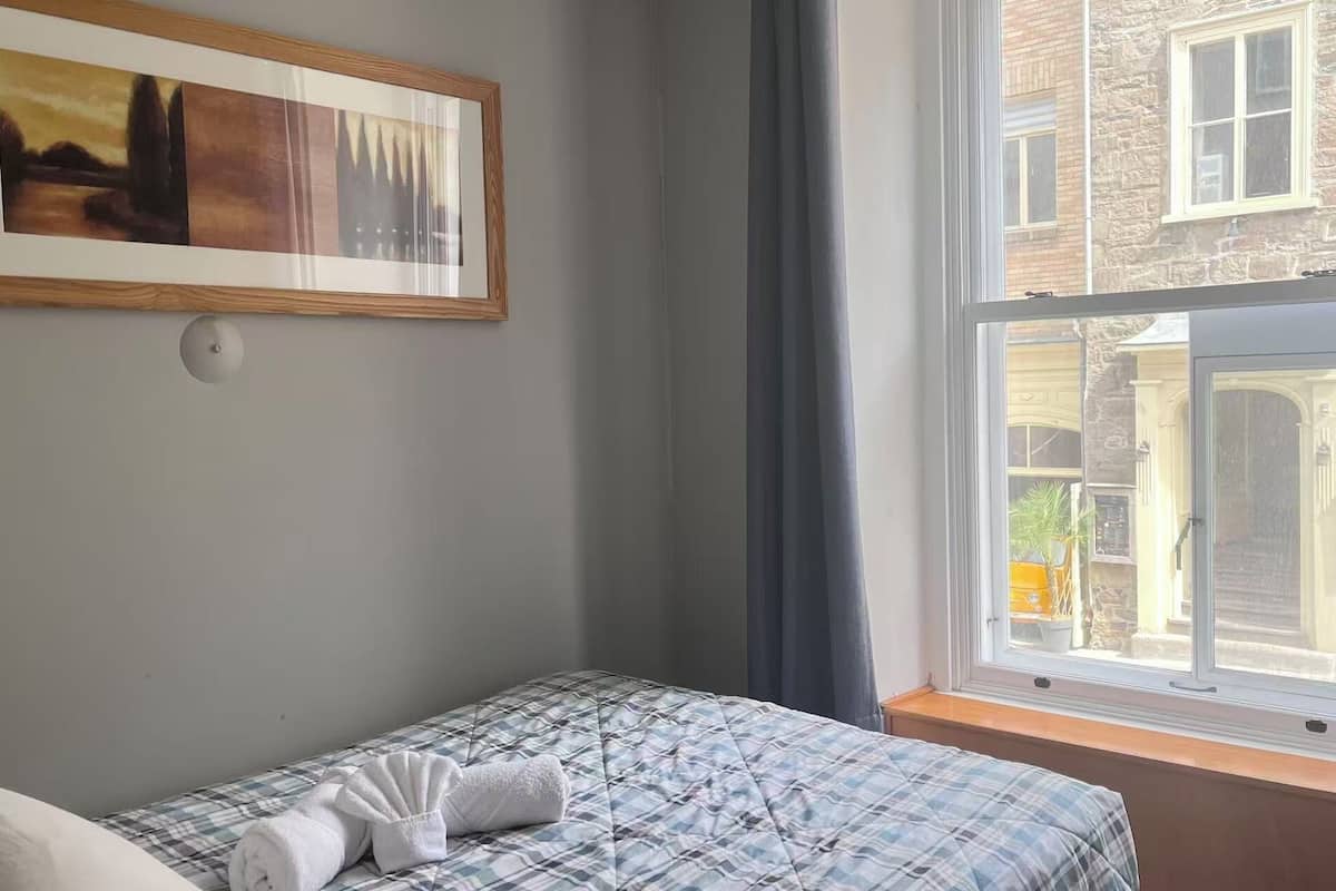 A hotel room with a bed, a painting on the wall, and a window with a view of buildings.