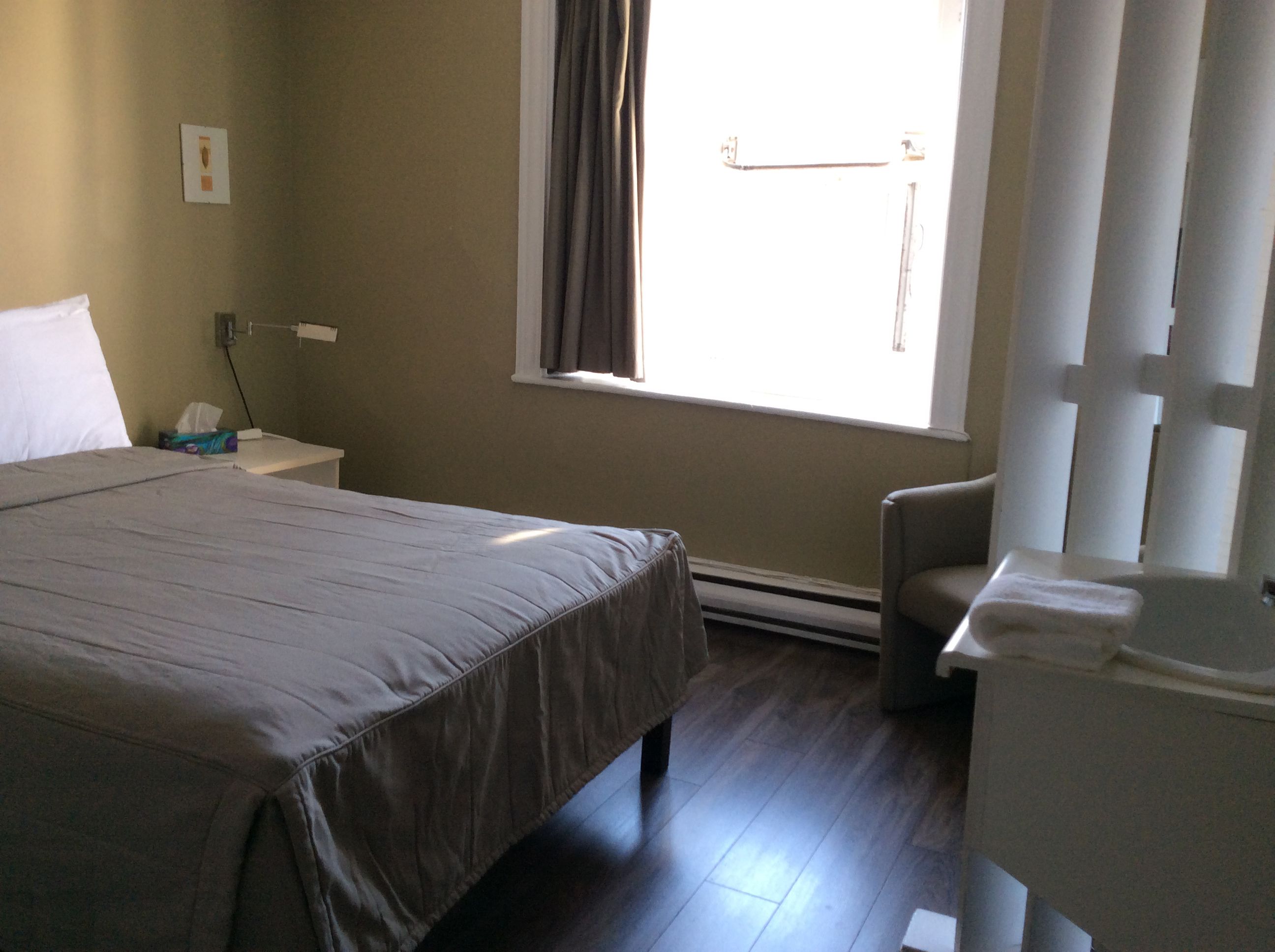 Une chambre d’hôtel avec un lit, une petite table de chevet, une chaise, une fenêtre avec des rideaux et un radiateur.
