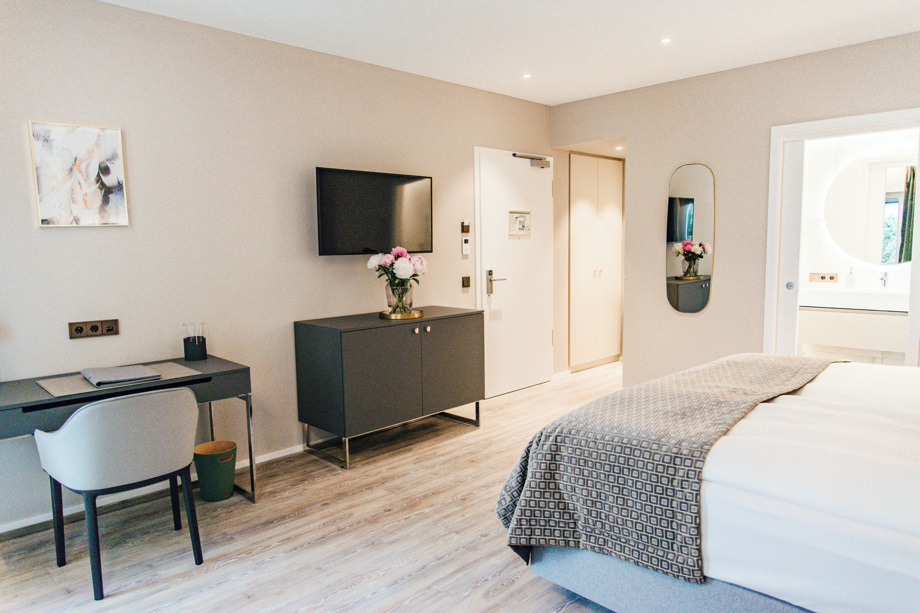 A modern hotel room with a bed, a desk with a chair, a television, a wardrobe, and a bathroom with a mirror.
