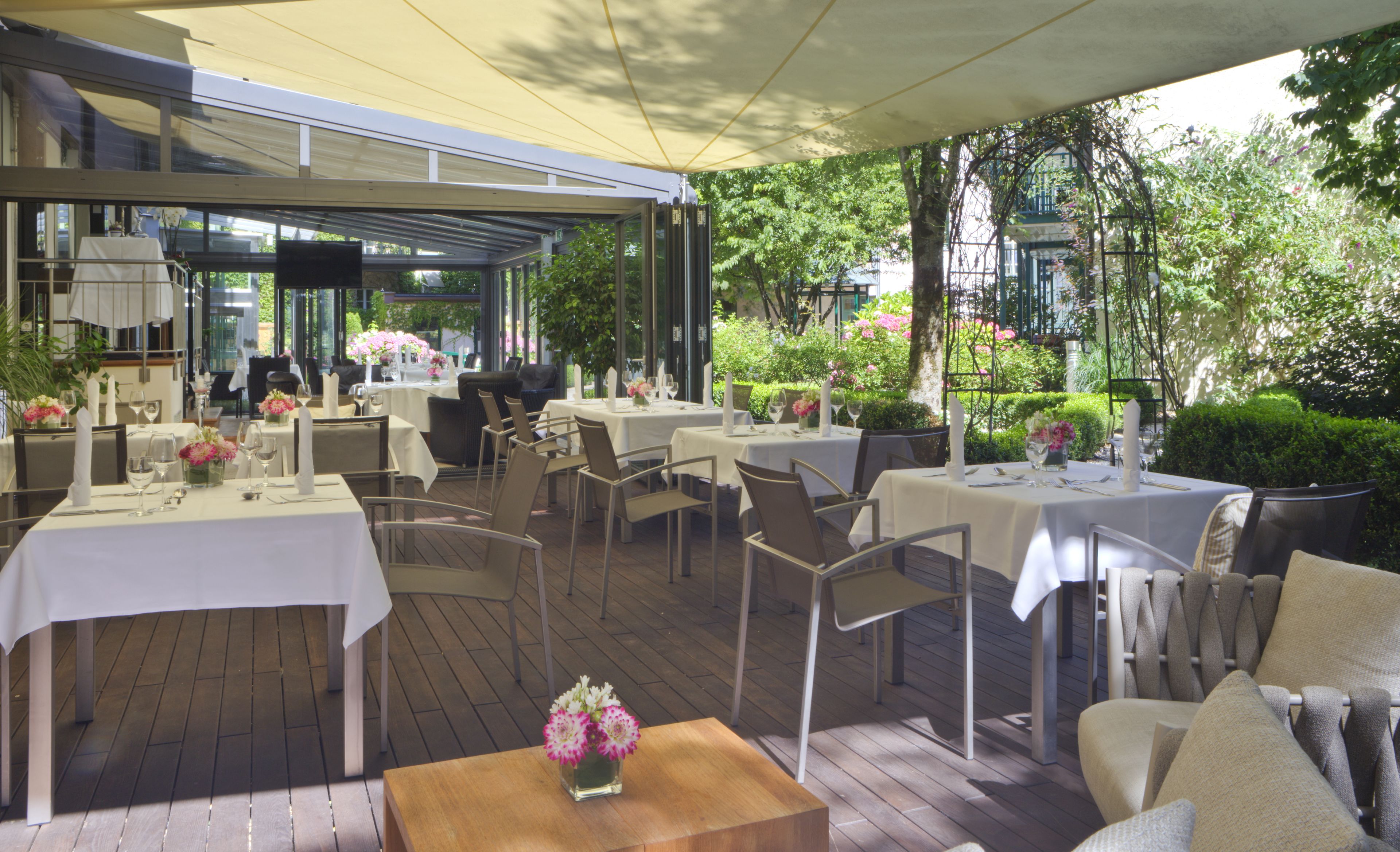 An outdoor dining area with tables and chairs, covered by a canopy, and surrounded by greenery.