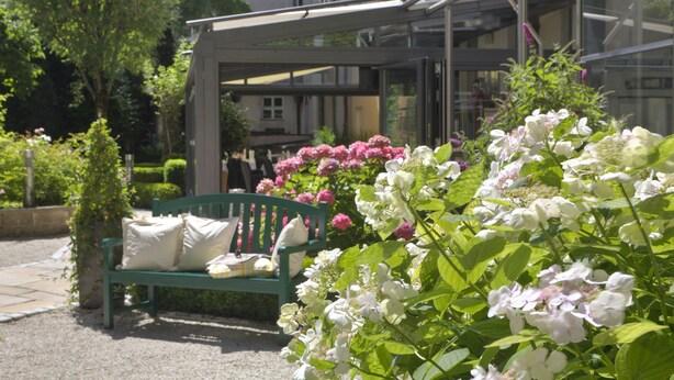 A green bench with cushions in a garden area with pink and white flowers, a glass structure, and a building in the background. - MAXIMILIAN MUNICH Apartments & Hotel  (Munich)