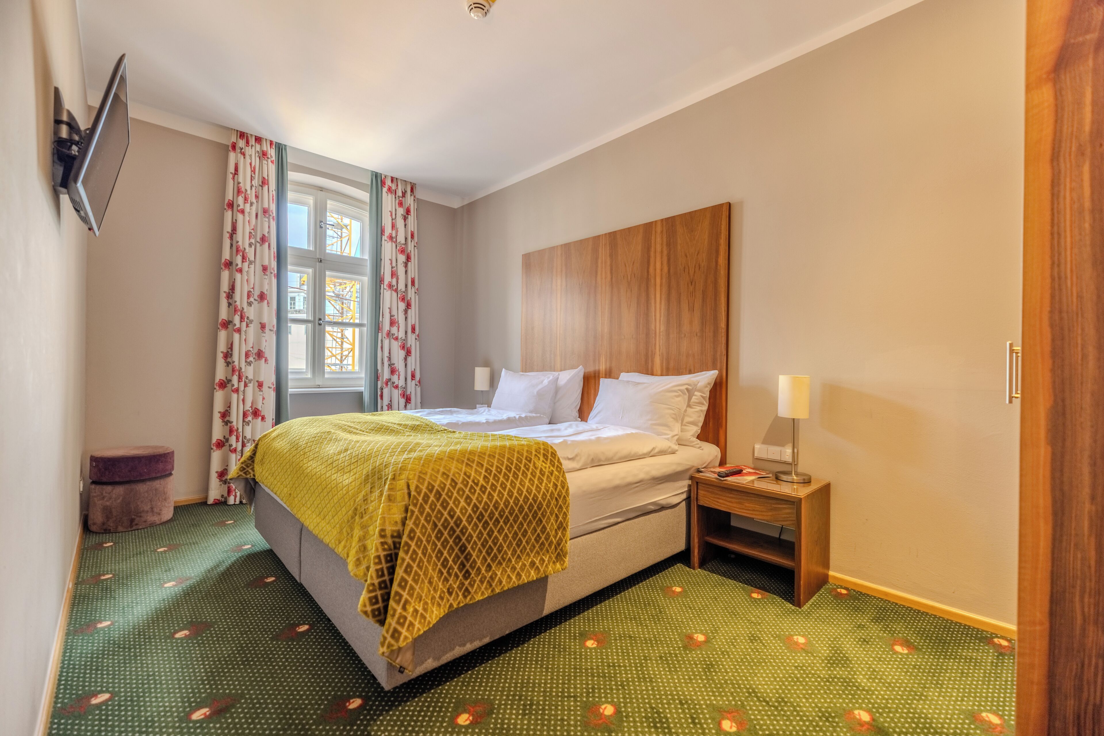 Quarto de hotel com cama, mesa de cabeceira, televisão na parede e janela com cortinas.
