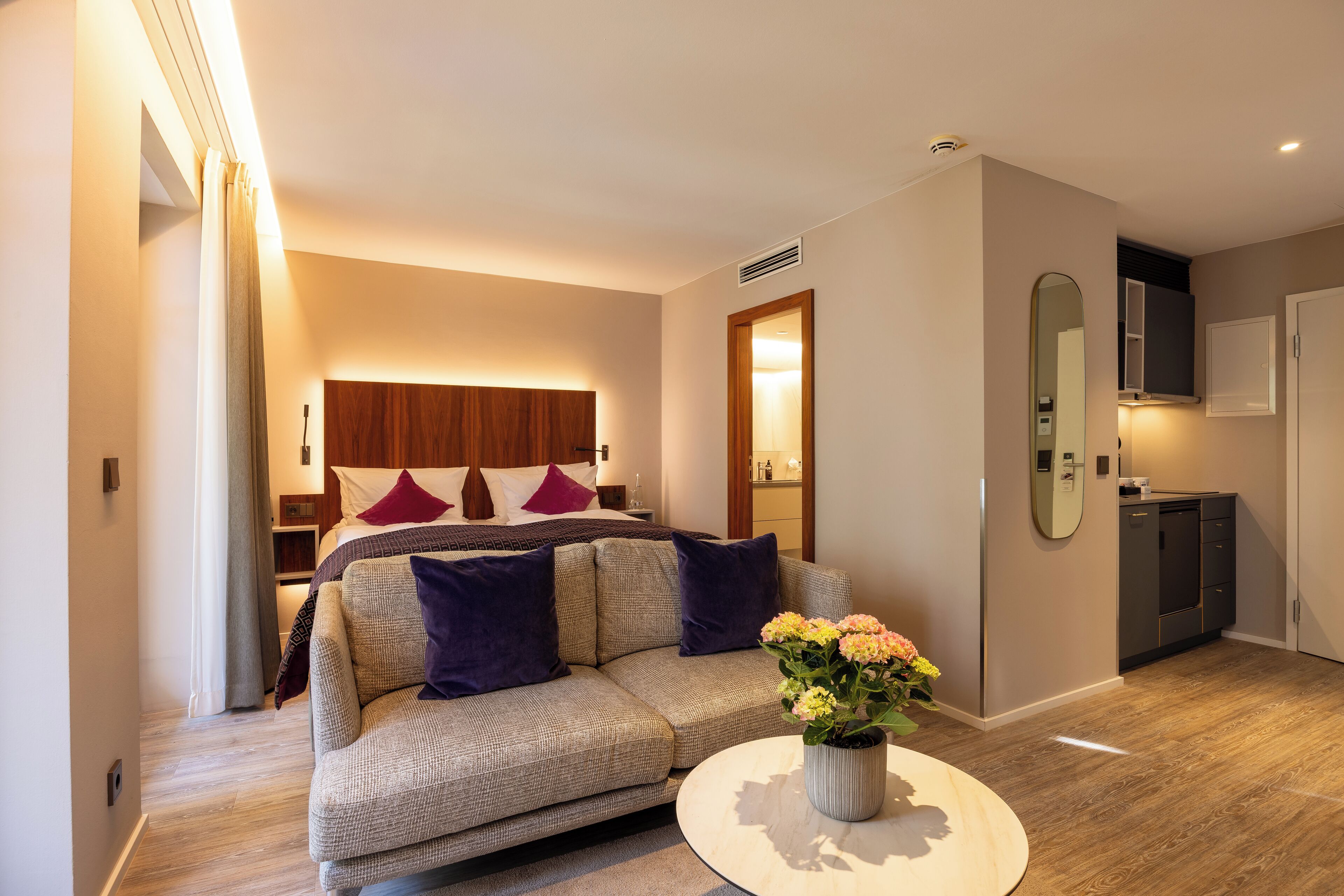 A modern hotel room with a sofa, a round coffee table with flowers, a bed with a wooden headboard, a mirror, and a small kitchenette.