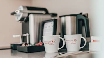 Dos tazas de café blancas con la marca 'Maximilian Munich' sobre una mesa, una cafetera y una caja de pasteles al fondo.