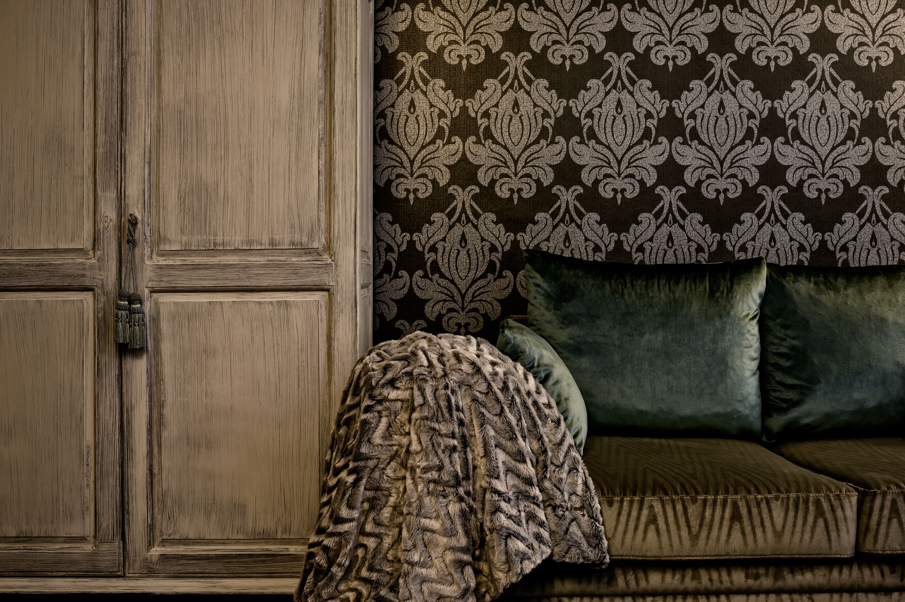 A room with a wooden wardrobe, a patterned wallpaper, and a green sofa with cushions.