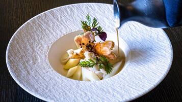Une assiette blanche à texture, sur laquelle est servi un plat de fruits de mer, comprenant une tête de poisson, agrémenté d’herbes, et une sauce est versée dessus.