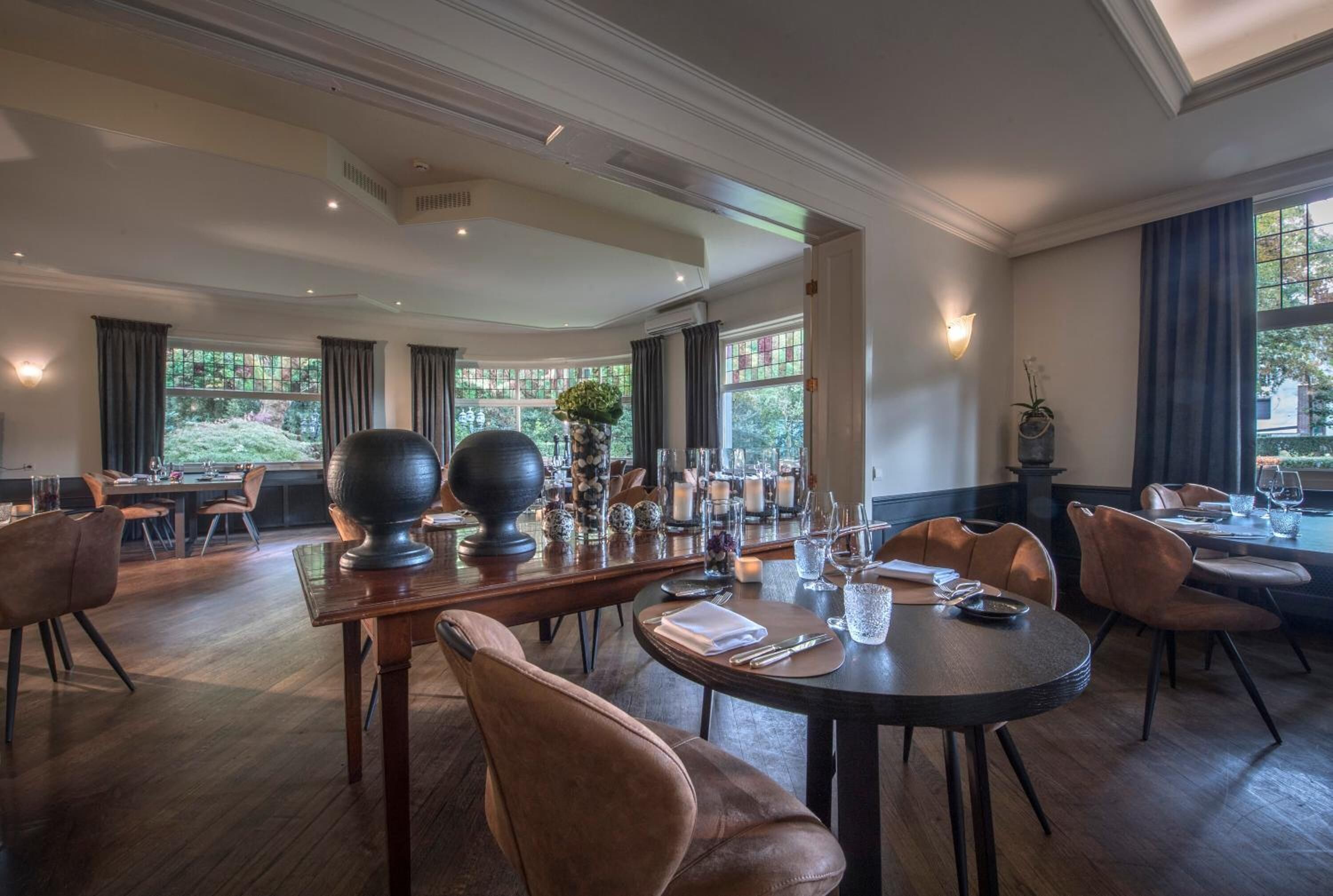 A spacious dining area with high ceilings, large windows, and a view of greenery outside. Tables are set with candles and glassware.