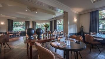 A spacious dining area with high ceilings, large windows, and a view of greenery outside. Tables are set with candles and glassware.
