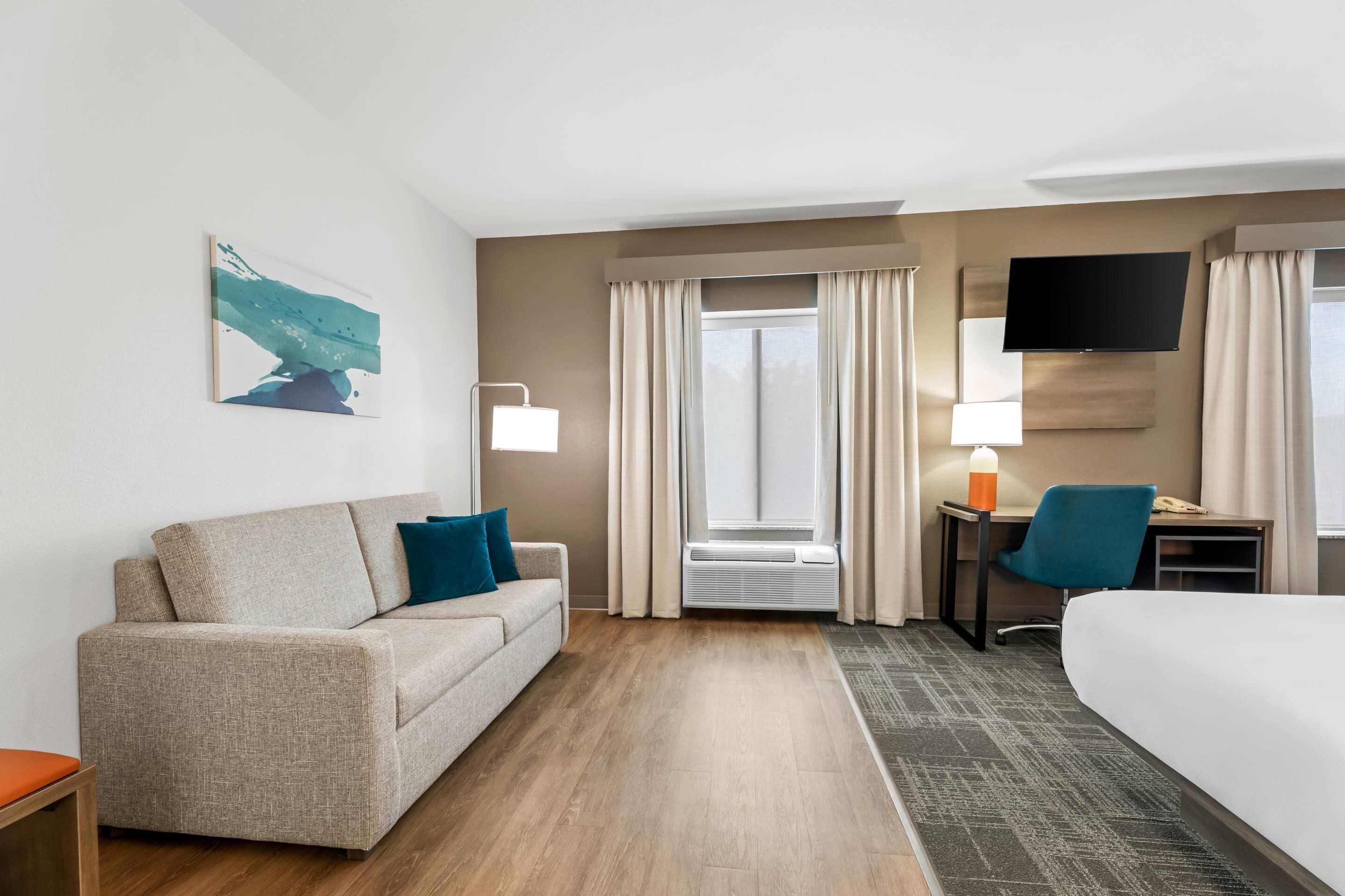 A hotel room with a sofa, a desk with a chair, a bed, a TV, and a window with curtains.