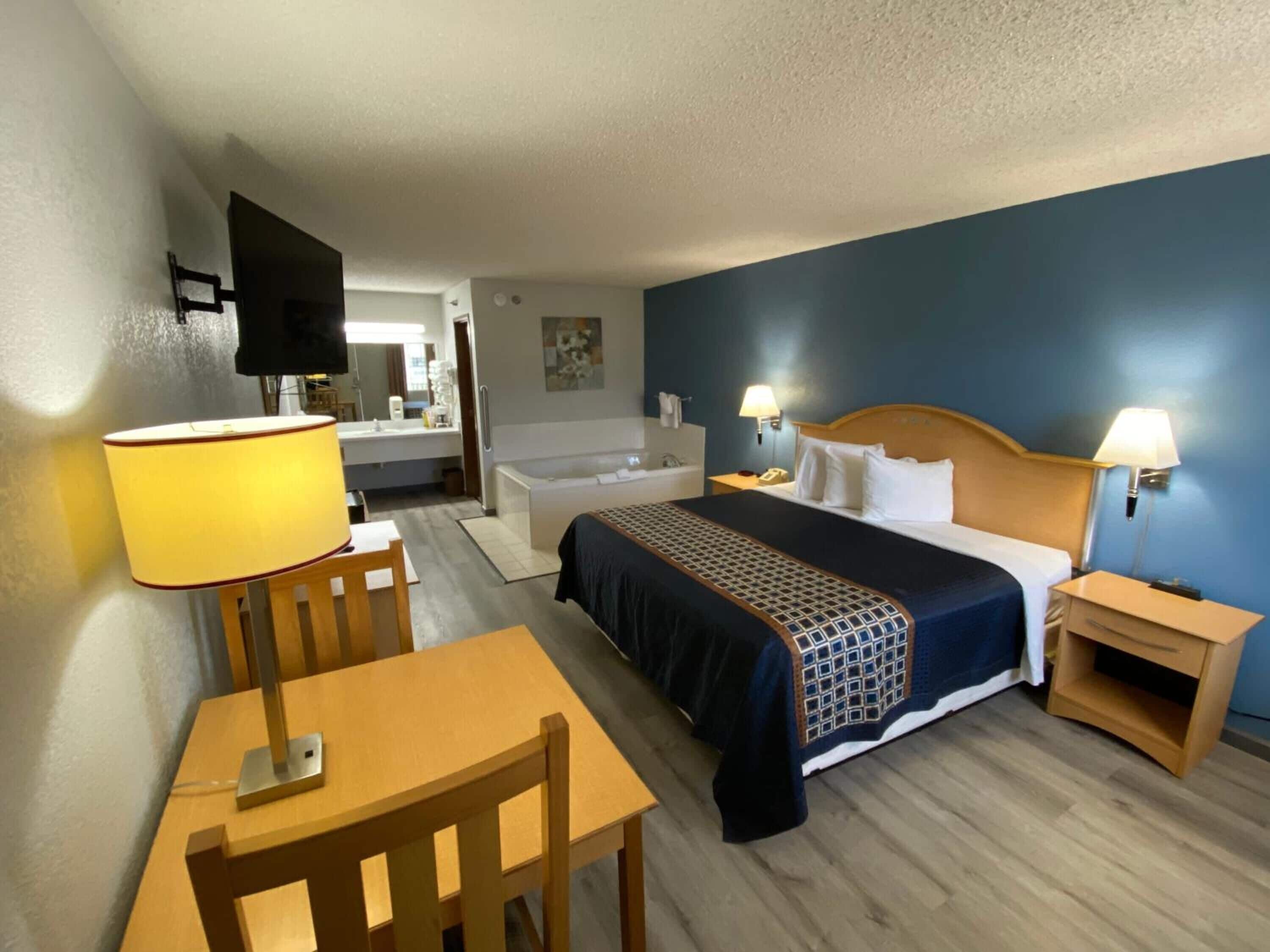 Habitación de hotel con cama, mesitas de noche, escritorio con silla, lámpara, televisor y baño con bañera y lavamanos.