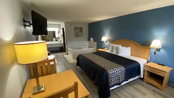 Habitación de hotel con cama, mesitas de noche, escritorio con silla, lámpara, televisor y baño con bañera y lavamanos.