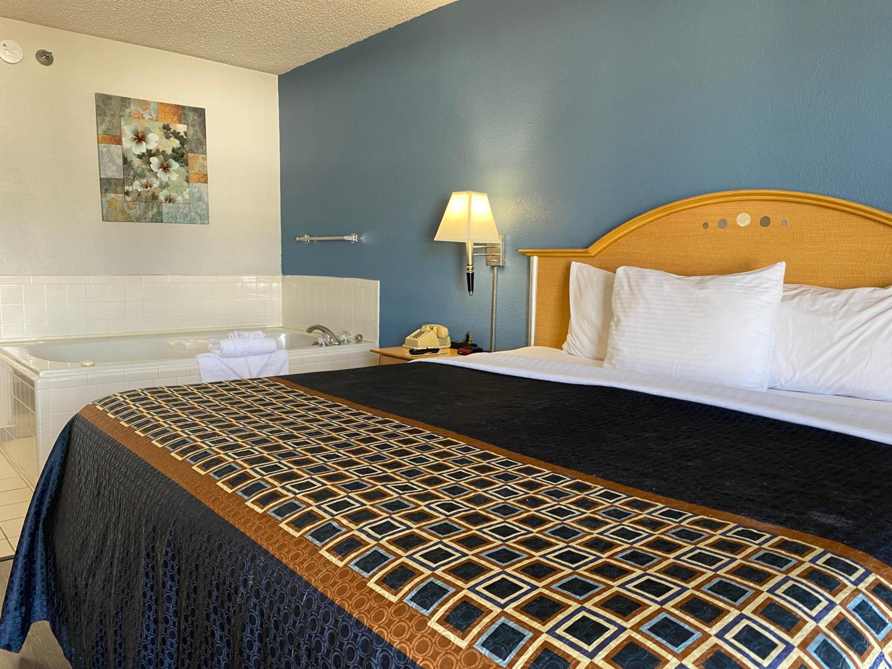 Habitación de hotel con una cama grande, una bañera, una mesita de noche con lámpara y un teléfono en la pared.