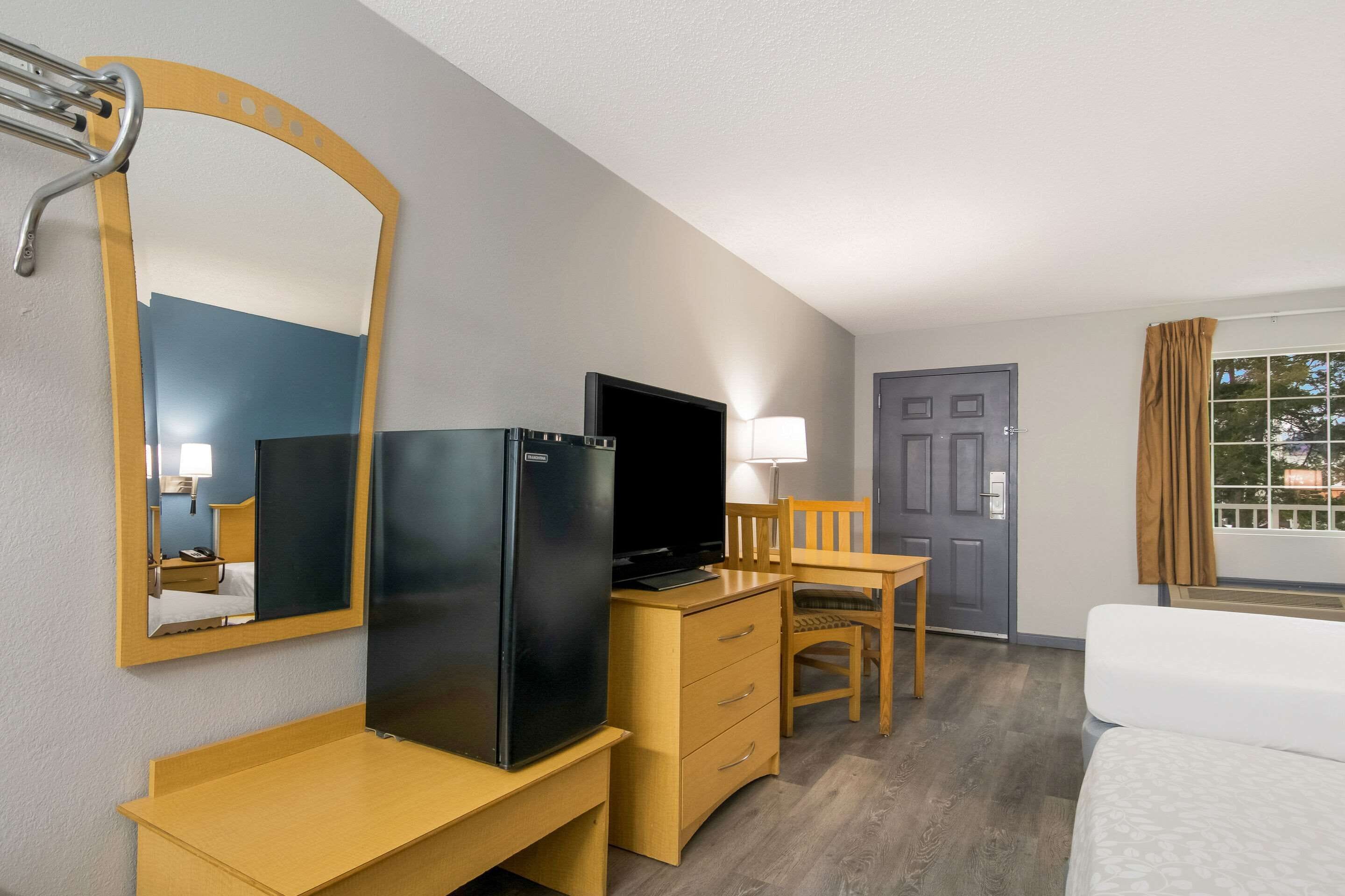 A hotel room with a bed, a desk with a television, a refrigerator, a chair, a lamp, and a window with curtains.